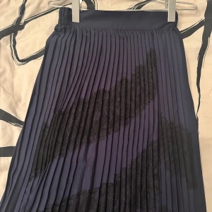 Plisserad mörkblå långkjol med spets - Snygg plisserad långkjol i mörkblått med svarta spetsdetaljer i diagonala mönster. Kjolen har hög midja och ett elegant fall. Perfekt för dig som vill ha en stilren men ändå unik look.