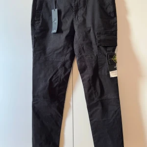 Svarta cargobyxor från Stone Island - Svarta cargobyxor från Stone Island med klassisk patch på benet. Byxorna har flera fickor, raka ben och är tillverkade i bomull. Snygg och funktionell design med coola detaljer och modern passform. Perfekta för dig som gillar streetwear och vill ha något extra.