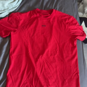 Röd t-shirt från Hugo Boss - Säljer en röd t-shirt från Hugo Boss med diskret logga på bröstet. Klassisk passform med rund hals och korta ärmar. Tillverkad i mjuk bomull som känns skön mot huden. Perfekt för dig som gillar stilrena och enkla plagg. Använd runt 2-3 gånger.