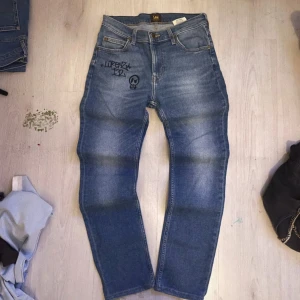 Blå raka jeans från Lee med tryck - Snygga blå jeans från Lee med raka ben och klassisk femficksdesign. Jeansen har ett unikt svart tryck framtill med text och döskalle, samt Lee-lapp bak i midjan. Tillverkade i slitstarkt denimtyg som ger en cool och avslappnad vibe.