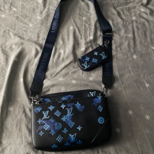 Louis Vuitton svart axelväska med blått mönster - Snygg svart axelväska från Louis Vuitton med blått monogrammönster och coola detaljer. Väskan har justerbar axelrem med logga och en mindre avtagbar ficka. Tillverkad i skinn och perfekt för dig som vill sticka ut med en unik design.