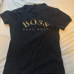 Svart Hugo Boss t-shirt med guldtryck - Svart t-shirt från Hugo Boss med stort BOSS-tryck i guld på bröstet och mindre logga i nacken. Klassisk passform och rund hals. Materialet är mjuk bomull som känns skönt mot huden. Perfekt för dig som gillar stilrena och lyxiga detaljer.