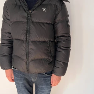 Svart pufferjacka från Calvin Klein - Svart pufferjacka från Calvin Klein Jeans med stor huva och dragkedja framtill. Jackan har logga på bröstet och ärmen, samt två fickor med dragkedja. Perfekt för kalla dagar och riktigt snygg till jeans.