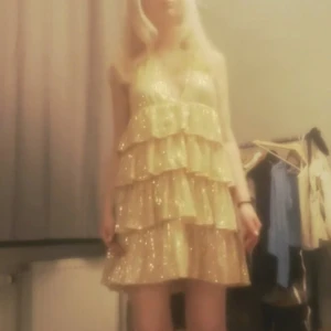 Glittrig guldig volangklänning - Supersöt kort klänning i guld med glittriga paljetter och flera volanglager. Djup v-ringning och öppen rygg ger en extra snygg look. Perfekt för dig som vill sticka ut och glänsa! Klänningen är ärmlös och har en figurnära passform upptill.