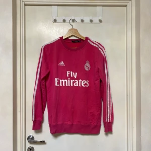Rosa Real Madrid tröja från Adidas - Snygg rosa långärmad fotbollströja från Adidas med Real Madrid-logga och Fly Emirates-tryck på bröstet. Klassiska vita ränder längs ärmarna och ribbad hals samt muddar. Materialet är mjukt och bekvämt, perfekt för dig som gillar sportig stil.