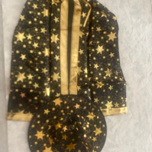 Svart trollkarlshatt med guldstjärnor - Cool svart trollkarlshatt med glittriga guldstjärnor och matchande cape. Perfekt för maskerad eller temafest. Hatten har klassisk spetsig form och är tillverkad i lätt syntetmaterial med metallic-detaljer.