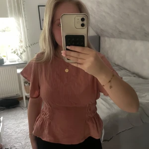 Korall  blus - Superfin korall blus med korta volangärmar och smockdetaljer i midjan. Blusen har en lätt rynkad struktur och knäppning bak i nacken. Perfekt till jeans eller kjol för en söt och trendig look.