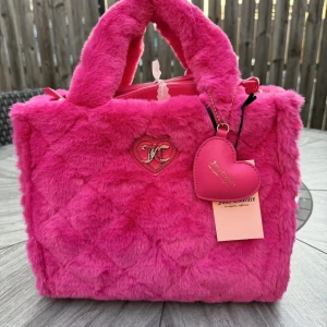 Rosa fluffig handväska Juicy Couture - Superfin rosa handväska från Juicy Couture i mjuk fuskpäls med hjärtformat hänge och JC-logga i silver. Väskan har både handtag och axelrem, samt dragkedja upptill. Perfekt för dig som vill sticka ut med en färgstark accessoar.