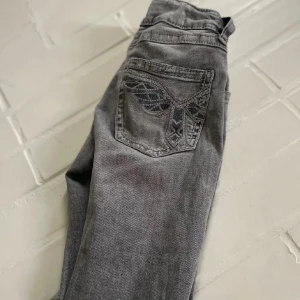 Grå jeans med broderade bakfickor Gina Tricot - Snygga grå jeans från Gina Tricot i storlek 158. Jeansen har broderade detaljer på bakfickorna och en cool knapp framtill. Modellen är rak med klassisk passform och tillverkad i mjukt denimtyg. Perfekta för dig som gillar en stilren men ändå unik look.
