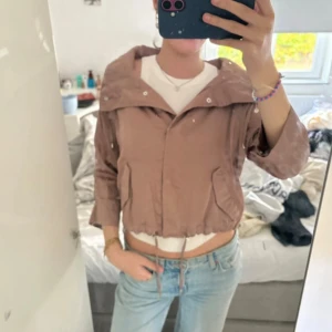 Beige cropped vindjacka med snörning - Säljer en beige cropped vindjacka med bred krage och snörning i midjan. Jackan har trekvartsärmar och två fickor framtill. Perfekt för dig som gillar en avslappnad och trendig stil.