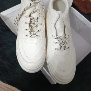 Vita sneakers med snörning - Snygga vita sneakers med rund tå och platt sula. Skorna har kontrasterande snören med svart mönster och är tillverkade i syntetmaterial. Perfekta för dig som gillar stilrena och fräscha skor med modern vibe.