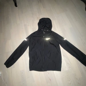 Svart Nike vindjacka med huva - Svart Nike Running vindjacka med huva och reflekterande logga på bröstet. Jackan har hel dragkedja framtill och är tillverkad i lätt polyester, perfekt för löpning eller utomhusaktiviteter. Snygg och sportig design med lång ärm.
