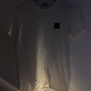 Vit t-shirt från Stone Island - Vit t-shirt från Stone Island med klassisk rund hals och korta ärmar. På bröstet sitter den ikoniska svarta patchen med logga. T-shirten är i mjuk bomull och har en clean, stilren look som passar perfekt till jeans eller shorts.