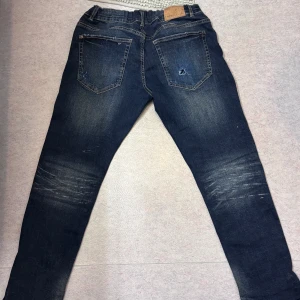 Mörk blåa jeans med snygg slitning  - Säljer ett par mörk blåa jeans i mörkblå tvätt med snygga slitningar och whiskers på låren. Modellen är mellan regular och slim fit och har klassiska fem fickor, samt en patch baktill. Jag är 170 och väger 60kg