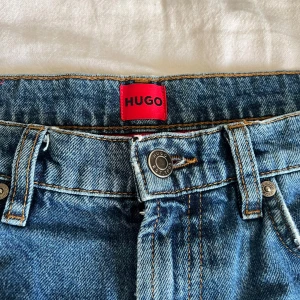 Blå slitna jeans från Hugo Boss - Säljer ett par blå jeans från Hugo Boss med coola slitningar och trasiga detaljer på benen. Klassisk femficksmodell med röd Hugo-label bak och fram. Jeansen har normal passform och är tillverkade i bomull för en skön känsla.