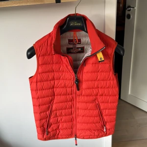 Röd dunväst från Parajumpers XS - Snygg röd dunväst från Parajumpers i storlek XS. Västen är quiltad med dragkedja framtill, två fickor med dragkedja och en gul detalj vid kragen. Insidan är ljusgrå och har flera praktiska innerfickor. Perfekt för lager-på-lager under kyliga dagar.