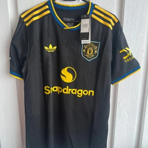 Manchester United svart matchtröja Adidas - Snygg svart Manchester United fotbollströja från Adidas med gula detaljer, blå inslag och tryck. Klassisk klubbmärke, sponsorlogga och nummer 10 Cunha på ryggen. Kortärmad modell i lätt funktionsmaterial, perfekt för match eller träning.