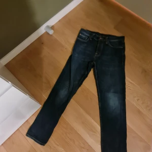 Mörkblå jeans från Nudie Jeans - Säljer ett par mörkblå jeans från Nudie Jeans med klassiska orange sömmar och snygga slitningar. Jeansen har rak passform, fem fickor och detaljer som logga på bakfickan och läderpatch i midjan. Perfekta för dig som gillar stilrena och tidlösa jeans.w29 L 30
