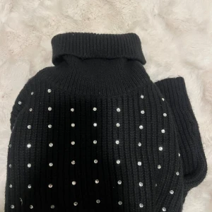 Svart polotröja med pärlor New Yorker - Ribbstickad svart polotröja från New Yorker dekorerad med glittrande pärlor över hela tröjan. Hög krage och långa ärmar ger en snygg och trendig look. Perfekt för dig som vill sticka ut lite extra. Aldrig använd