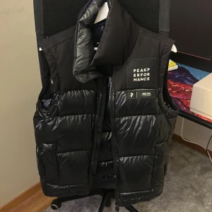 Svart dunväst Peak Performance M - Svart dunväst från Peak Performance med hög krage och tydlig logotyp på bröstet. Västen har quiltad design, dragkedja framtill och praktiska fickor. Materialet är glansigt och känns riktigt sportigt. Perfekt för lager-på-lager under kyliga dagar.