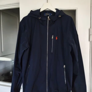 Marinblå vindjacka från Polo Ralph Lauren - Marinblå vindjacka från Polo Ralph Lauren med huva, dragkedja framtill och två sidofickor med dragkedja. Klassisk röd broderad logga på bröstet. Jackan har en stilren look och är tillverkad i lätt polyester, perfekt för blåsiga dagar.