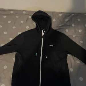 Svart hoodie från BOSS med dragkedja - Svart hoodie från BOSS med vit dragkedja och snörning i huvan. Diskret BOSS-logga på bröstet och coolt BOSS-tryck längs nederkanten på baksidan. Mjuk och skön, perfekt för chill eller streetstyle.