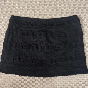 Svart spetskjol med volangdetaljer - Aldrig använt, köpt i Spanien. Supersnygg svart kortkjol med spets och flera volangrader runtom. Kjolen har en tight passform och är perfekt för dig som gillar en feminin och trendig look. Materialet är mjukt och följsamt, och spetsen ger en extra touch av stil.