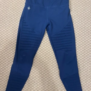 Blå leggings från Workout Empire XS - Snygga blå leggings från Workout Empire i storlek XS. De har ribbade detaljer längs benen och hög midja för en schysst passform. Perfekta för träning eller chill, tillverkade i stretchigt syntetmaterial som sitter skönt hela dagen.