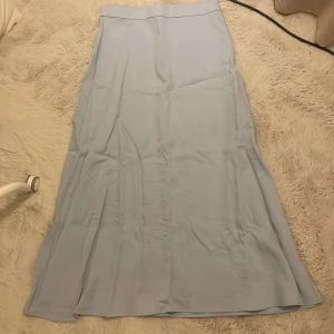 Ljusblå långkjol i mjukt tyg - Säljer en stilren ljusblå långkjol med enkel design och mjukt, följsamt tyg. Kjolen har en rak och flowy siluett som ger en chill vibe. Perfekt att matcha med sneakers eller sandaler för en clean look. Helt ny