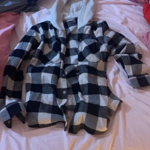 Svartvit rutig overshirt från Lindex - Overshirt från Lindex i storlek 164 med svartvitt rutigt mönster och mjuk grå huva. Skjortan har långa ärmar, två bröstfickor med lock och knappar framtill. Perfekt för lager-på-lager och chill stil. Materialet är bomull, vilket gör den skön att bära.