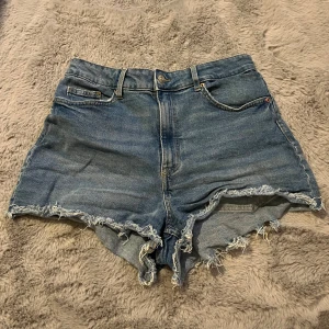 Blå jeansshorts från H&M Divided - Säljer ett par blå jeansshorts från H&M Divided i storlek 40. De är högmidjade med råa, fransiga kanter och klassisk femficksdesign. Perfekta för varma dagar och har en avslappnad, loose fit. Gjorda i bomull för en skön känsla.