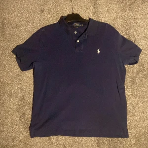 Mörkblå pikétröja Polo Ralph Lauren XL - Klassisk mörkblå pikétröja från Polo Ralph Lauren i stretch mesh-bomull. Tröjan har korta ärmar, två knappar vid kragen och den ikoniska broderade loggan på bröstet. Perfekt för en clean och stilren look.