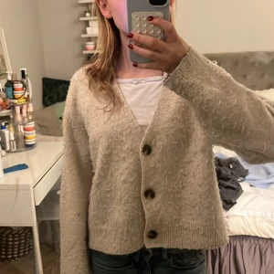 Beige kofta från Zara med knappar - Supermysig beige kofta från Zara med stora bruna knappar framtill. Koftan är stickad och har en lite fluffig yta, v-ringad och med långa ärmar. Perfekt att slänga på sig när det är kyligt ute.