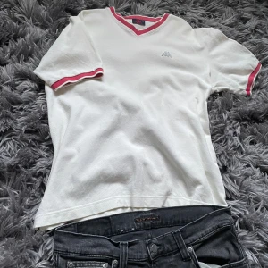 Kappa - Vit piké från Kappa med v-ringning och röda ribbade kanter vid hals och ärmslut. Klassisk broderad Kappa-logga på bröstet och skön passform.