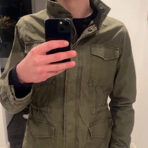 Olivgrön parkajacka med fickor - Säljer en olivgrön parkajacka med fyra stora fickor framtill och hög krage. Jackan har dragkedja och knappar, samt justerbar midja för snygg passform. Perfekt för dig som gillar en stilren och avslappnad look. Sitter perfekt om du är mellan 180-185