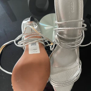 Silvriga klackskor från ASOS Design - Snygga silvriga klackskor från ASOS Design med spetsig tå och tunna remmar som snöras runt vristen. Skorna har hög klack och är tillverkade i syntetmaterial med metallicfinish. Perfekta för att sticka ut och ge en futuristisk touch till din outfit. Helt ny oanvänd