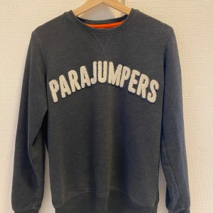 Mörkgrå fleece-tröja Parajumpers - Mörkgrå långärmad fleece-tröja från Parajumpers med stor vit logga framtill och patch på ärmen. Tröjan har rund halsringning, ribbade muddar och orange detalj i nacken. Perfekt för kyliga dagar.