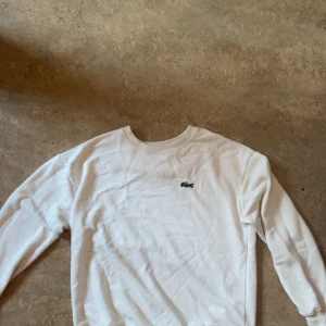 Vit sweatshirt från Lacoste - Vit sweatshirt från Lacoste med klassisk rund hals och broderad grön krokodil på bröstet. Tröjan har ribbade muddar och är tillverkad i mjuk bomull som är skön att bära. Perfekt för dig som gillar stilrena och sportiga plagg med ikonisk logga.