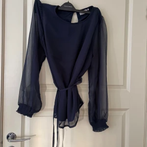 Marinblå blus med skärp - Snygg marinblå blus med transparenta långärmar och rund hals. Blusen har ett knytband i midjan för att skapa en omlott-effekt och ett elegant fall. Perfekt för dig som gillar stilrena och moderna plagg med lite extra detaljer.