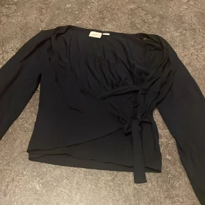 Mörkblå kofta från Gina Tricot - Säljer en mörkblå kofta från Gina Tricot med långa ärmar och snygg knytning i sidan. Toppen har en v-ringad design och är tillverkad i mjukt, lätt polyester-material som sitter bekvämt och ger en stilren look.