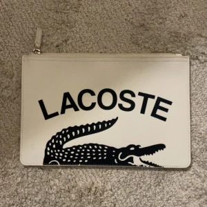 Vit kuvertväska/nässecer från Lacoste - Snygg vit kuvertväska/nässecer från Lacoste med stort svart krokodilmotiv och logga på framsidan. Väskan har dragkedja upptill. Aldrig använd
