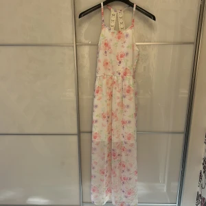 Blommig långklänning med spetsdetalj - Säljer en romantisk långklänning med blommigt mönster i pastellfärger. Klänningen har smala axelband och en snygg spetsdetalj i ryggen. Materialet är lätt och luftigt, perfekt för varma dagar. Använt 1 gång på avslutning i 6:an!