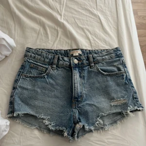 Blå jeansshorts från Gina Tricot - Snygga blå jeansshorts från Gina Tricot x Iza & Elle med råa kanter och slitningar framtill. Klassisk femficksmodell med knapp och dragkedja. Perfekta för sommaren och ger en avslappnad vibe.