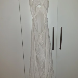 Vit långklänning från New Look - Superfin vit långklänning från New Look med v-ringad front, volangdetaljer på axelbanden och snygg öppen rygg. Klänningen har ett diskret rutigt mönster och är tillverkad i bomull, vilket gör den lätt och skön. Perfekt för varma dagar och har en loose passform.