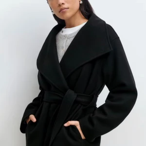 Svart kort jacka  Gina Tricot stl S - Snygg svart kort kappa med bred krage och omlottmodell, knyts med bälte i midjan. Långärmad och med fickor på sidorna. Endast provad ett par gånger. Ångrade köpet. Helt ny utan lappar. Stl small Nypris 700kr 