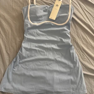 Ljusblå  klänning från Alo Yoga - Supersöt ljusblå klänning från Alo Yoga med vita kontrastdetaljer längs kanterna och tunna axelband. Klänningen har en figurnära passform och är tillverkad i mjukt, stretchigt syntetmaterial som känns skönt mot huden. Perfekt för dig som gillar sportig och fräsch stil. Klänningen är i storlek L men funkar även för någon som är i storlek M dessutom är det inbyggda shorts i