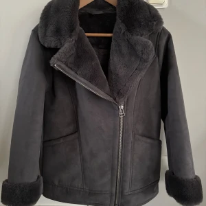 Mörkgrå pälsjacka med dragkedja - Snygg mörkgrå pälsjacka med fluffigt foder och bred krage. Jackan har en silvrig dragkedja framtill och uppvikta ärmslut med pälsdetaljer. Perfekt för kalla dagar 