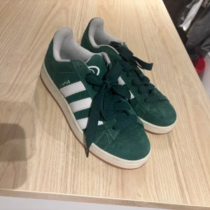 Adidas Campus gröna sneakers - Säljer ett par Adidas Campus sneakers i mörkgrön mocka med klassiska vita ränder på sidorna och vit logga på plösen. Skorna har rund tå, platt sula och snörning. Insidan är ljusgrå och yttersulan är beige med brun kant. Perfekt för dig som gillar retrostil och streetwear.