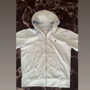 Vit hoodie från Polo Ralph Lauren - Snygg vit hoodie från Polo Ralph Lauren med dragkedja och huva. Klassisk logga broderad på bröstet, två stora fickor framtill och snörning i huvan. Perfekt för en chill och stilren look.