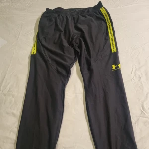 Svarta träningsbyxor Under Armour L - Svarta träningsbyxor från Under Armour med gula detaljer och logga på benet. Byxorna har elastisk midja med snörning och gula band längs sidorna med text. Fast pris 300 kr.